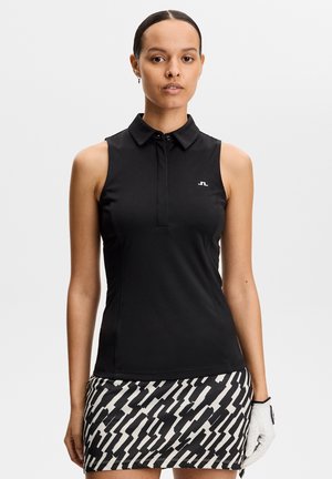 Frau trägt ein ärmelloses schwarzes Hemd mit Kragen und einen schwarz-weiß gemusterten Rock, hält einen weißen Golfhandschuh an ihrer linken Hand.