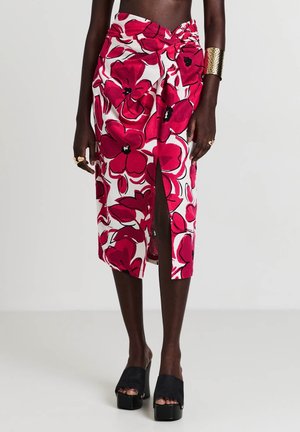 Mujer con una falda midi blanca con grandes flores rojas y una abertura frontal, combinada con zapatos negros de plataforma abiertos en la puntera y joyas de oro.