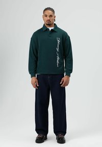 Donkergroen oversized poloshirt met lange mouwen, voorzien van witte schrifttekst aan de zijkant. Gecombineerd met donkerblauwe wijd uitlopende jeans en zwarte schoenen.