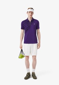 Paarse polo shirt met een ruitpatroon, witte shorts, groene schoenen en een witte visor. Houdt een netzak vast met groene tennisballen.