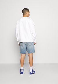 Witte sweatshirt met een relaxte pasvorm, gecombineerd met lichtblauwe denimshorts. Model draagt blauwe hoge sneakers met een witte zool en branding.