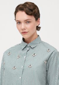 Stripet grønn og hvit skjorte med knapper, prydet med sølvblomsterdekorasjoner, en spiss krage og et figursydd design.
