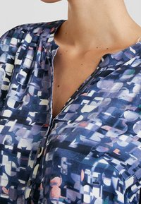 Koszula z guzikami w niebieskim wzorze z abstrakcyjnymi kształtami i kolorami. Gładki materiał z kołnierzykiem i delikatnymi guzikami. Krótkie rękawy.