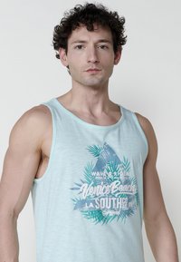 Svetlozeleni top bez rukava iz lahkog materijala sa grafičkim dizajnom "Venice Beach" uz tropske akcenta i trouglastim uzorkom.
