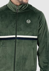 Giacca zip-up in velluto verde con collo alto, strisce orizzontali bianche e navy, tasche laterali e logo circolare sul petto.