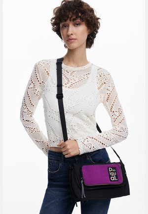 Young woman wearing a white crochet long-sleeve top and jeans, holding a black and purple Desigual crossbody bag with adjustable strap.