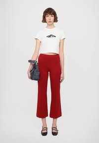 T-shirt blanche courte avec impression noire, associée à un pantalon large rouge. Le mannequin tient un sac gris foncé avec plusieurs poches, portant des chaussures noires.