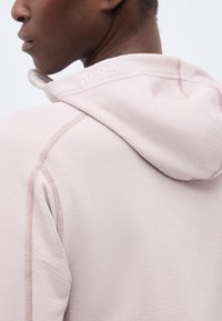 Inimene, kes kannab heledat roosat kapuutsiga dressipluusi, millel on kaela lähedal seljal vasakul küljel nähtav "Houdini" logoga tikand.