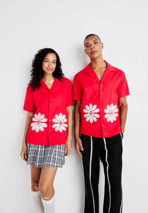 Rode shirts met korte mouwen en witte bloemen borduurwerk aan de zoom, gedragen met een geruite rok en zwarte broeken met witte accenten.