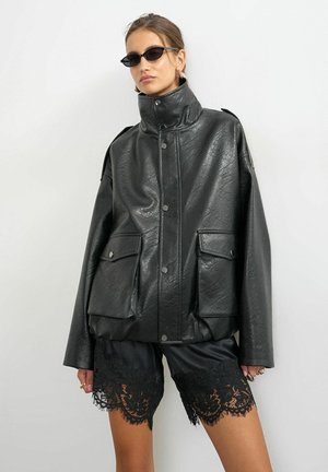 Mulher vestindo um casaco de couro preto oversized com gola alta e grandes bolsos frontais, calções pretos com acabamento em renda e pequenos óculos de sol pretos.