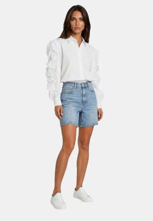 Donna in piedi frontale che indossa una camicia bianca a volant a maniche lunghe, shorts di jeans blu e sneakers bianche su uno sfondo semplice.