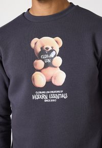 Tamno siva dukserica s grafikom svijetlosmeđeg medvjedića koji drži crno srce s natpisom "CLOSURE LDN" i bijelim sloganom branda ispod.
