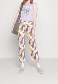 Jasnofioletowy top bez rękawów z graficznym nadrukiem; krótki, związany w talii. Brązowe, żółte i białe spodnie o szerokich nogawkach z wzorem; szara torba; beżowe buty wsuwane.