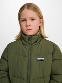 Blondes Kind mit glatten Haaren trägt eine grüne, gesteppte hummel Sportswear-Jacke mit Reißverschluss und schaut mit neutralem Gesichtsausdruck geradeaus.