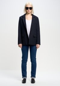 Svart rutigt kavaj med snibbade slag, kombinerat med en vit topp och figurnära blå jeans. Modellen bär mörka solglasögon och svarta slip-on skor.