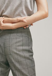 Pantalon à carreaux noirs et blancs avec une coupe ajustée, comprenant des poches et une texture de tissu lisse. Porté avec un t-shirt gris.