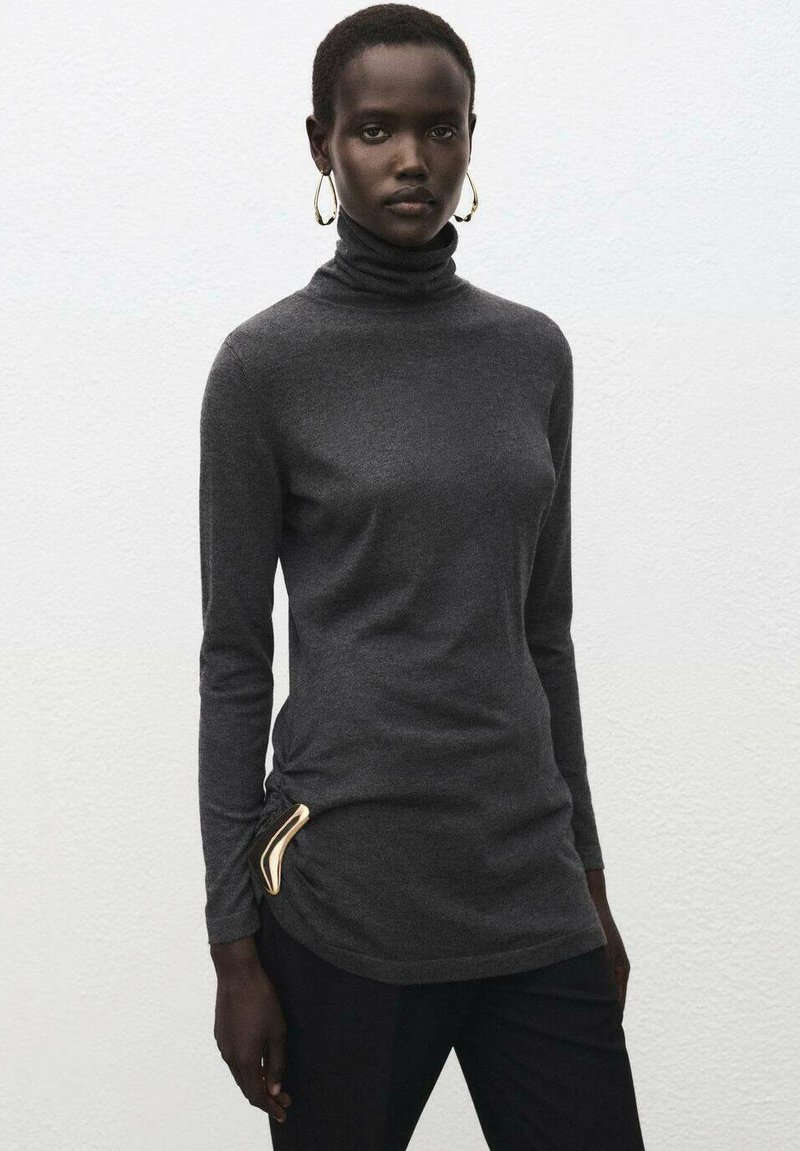 Suéter de cuello alto gris en una tela suave con mangas largas, que presenta un detalle dorado en el lateral. Combinado con pantalones negros.