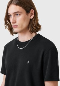 Camiseta de textura de gofrado negro con cuello redondo, que presenta un pequeño bordado de un cr�áneo de cabra blanco en el lado izquierdo del pecho. Collar de cadena de plata visible.