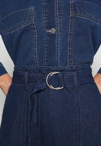 Vestido de mezclilla con mangas largas, de material azul, que cuenta con una cintura ceñida con cinturón y herrajes plateados en forma de D. Dos bolsillos en el pecho con costuras visibles.