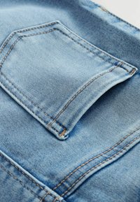 Tissu en denim bleu clair avec une poche à rabat visible. Des coutures contrastées entourent la poche et longent les bords, mettant en valeur une texture lisse.