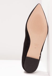 Chaussure à talon haut en suede noir avec un bout pointu, une silhouette élancée et une semelle en caoutchouc texturé. Les marquages incluent "FABRIQUÉ EN ESPAGNE" et la taille 38.