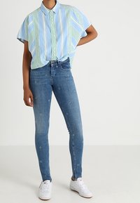 Ljusblåt skjorta med gröna och vita ränder, kombinerad med blå slim-fit jeans med subtila blommönster och ankellåsnar.