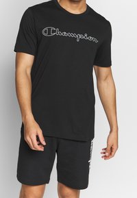 Man wearing a black Champion t-shirt and black shorts with white logo accents, standing against a plain light background.