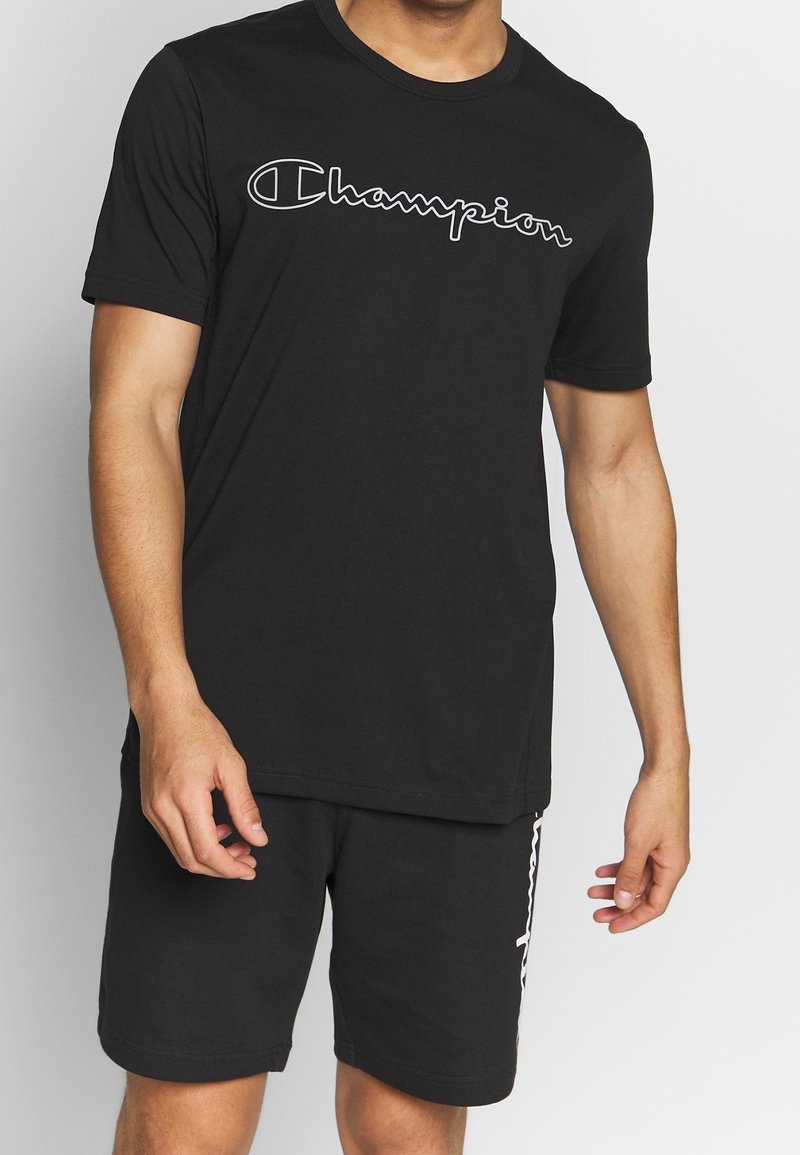 Man wearing a black Champion t-shirt and black shorts with white logo accents, standing against a plain light background.