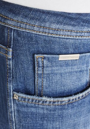 Bolsillo de jeans de mezclilla azul con costuras amarillas en contraste y una pequeña etiqueta metálica de la marca encima de la abertura del bolsillo.