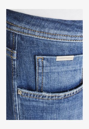 Bolsillo de jeans de mezclilla azul con costuras amarillas en contraste y una pequeña etiqueta metálica de la marca encima de la abertura del bolsillo.