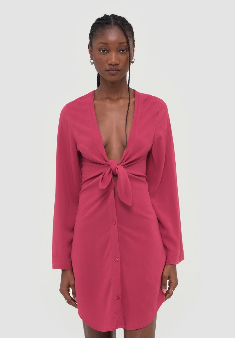 Femme aux cheveux tressés portant une robe rose à manches longues avec un profond décolleté en V, nouée devant, debout sur un fond clair uni.