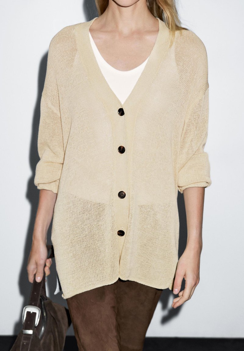 Kvinna som bär en beige stickad cardigan med svarta knappar över en vit topp, håller en mörkbrun mockaväska med en silverspänne.