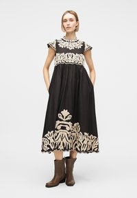 Žena v černých midi šatech s béžovou květinovou výšivkou a hnědými kotníkovými botami, stojící s rukama v kapsách na bílém pozadí.