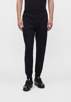 Partie inférieure du corps d'une personne portant un pantalon de jogging noir avec taille à cordon et des baskets noires brillantes sur un fond clair uni.