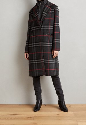 Personne portant un manteau à carreaux gris foncé longueur genoux avec des rayures rouges et gris clair, un pantalon noir et des bottines noires sur un sol en bois.