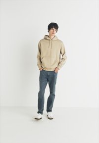 Sudadera beige con bolsillo frontal, combinada con jeans oscuros y zapatillas blancas. La tela parece suave, con un diseño simple y cordón ajustable.