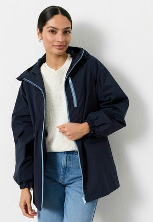 Frau trägt eine marineblaue Jacke mit hellblauen Reißverschlüssen über einem weißen Pullover und blauen Jeans, steht vor einer weißen, strukturierten Wand.