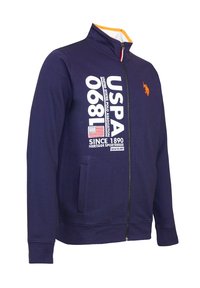 Marinblå zip-jacka med hög krage, orange inredning, vitt tryckt text och liten broderad logotyp. Har sidfickor och ribbade ärmslut.