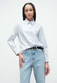 Cămașă cu dungi bleu deschis, cu nasturi, guler, mâneci lungi și un logo mic. Asortată cu blugi din denim deschis și o curea neagră.