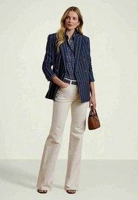 Naine kannab tumesinist triibulist bleiserit, mustrilist tumesinist särki, valgeid pükse ning hoiab pruuni kotti, seistes, üks käsi taskus.