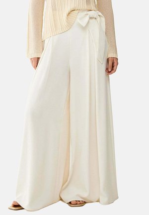 Pantaloni crema a gamba larga con cintura alta da annodare, abbinati a un maglione beige a maglia e sandali con punta quadrata visibili all'orlo.