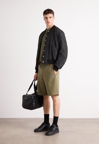 Svart bomberjakke over olivengrønn skjorte og shorts, kombinert med svarte joggesko og en svart duffelbag, med en glatt tekstur.