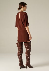 Abito corto marrone con maniche corte, caratterizzato da una apertura a chiave sulla schiena. Abbinato a stivali alti fino al ginocchio in pelle marrone con una texture morbida.