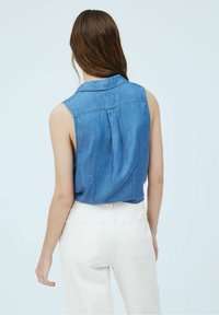 Jeansowa koszula bez rękawów z plisą z tyłu, w kolorze jasnoniebieskim, z kołnierzykiem, zestawiona z kremowymi spodniami.