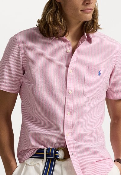 Košile s krátkým rukávem, na knoflíky, s růžovými a bílými vertikálními pruhy. Obsahuje náprsní kapsu a malou modrou nášivku na kapse.