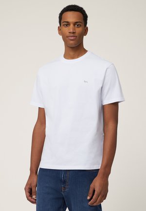Camiseta de algodón blanca con cuello redondo, mangas cortas y un pequeño logotipo bordado en el pecho. Se lleva con jeans azules.