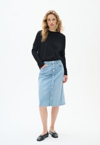 Haut noir à manches longues associé à une jupe crayon en denim bleu clair avec fermeture à boutons à l'avant, deux poches latérales et bords ourlés. Chaussures plates noires.