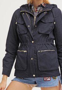 Navy blue hooded jacket with a cinched waist, zippered front, two chest pockets, and side pockets featuring silver hardware accents.