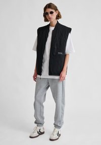 Schwarze ärmellose Weste mit einer Reißverschluss-Tasche, weißes T-Shirt, graue Jogginghose und beige Turnschuhe mit schwarzen und roten Akzenten, vor einem hellen Hintergrund.