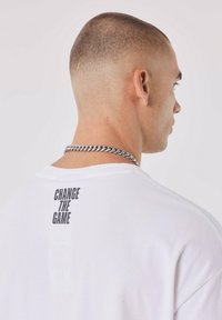 Witte katoenen T-shirt met een boxy fit, met daarop een zwarte print "CHANGE THE GAME" op de bovenrug. Zichtbare zilveren ketting.